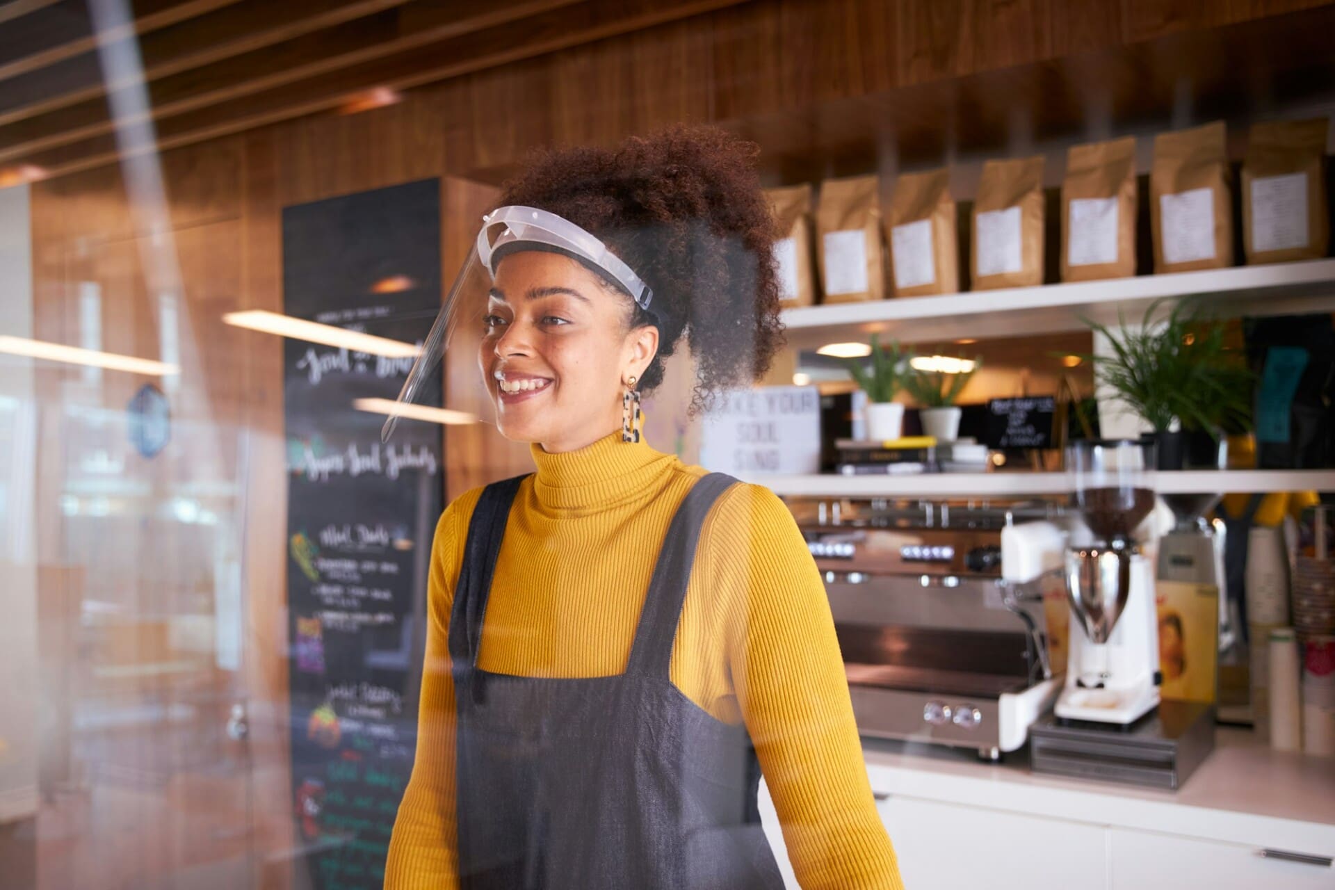 Female Small Business Owner Of Coffee Shop Wearing Face Shield Behind Counter During Health Pandemic